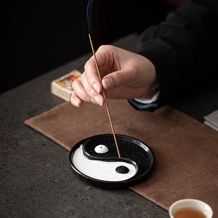 Hand placing incense stick into yin yang ceramic holder on home tea table with brown mat eye-level side view
