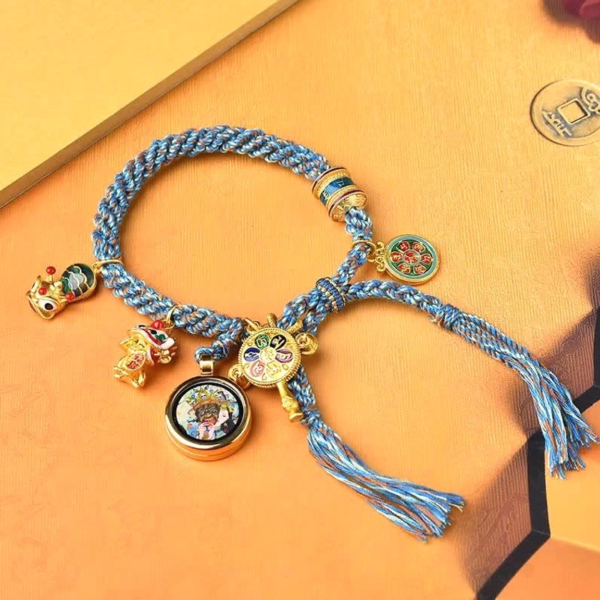 Zakiram Buddhist string bracelet flat lay featuring blue woven rope, enamel pendants, and decorative gold beads on orange background