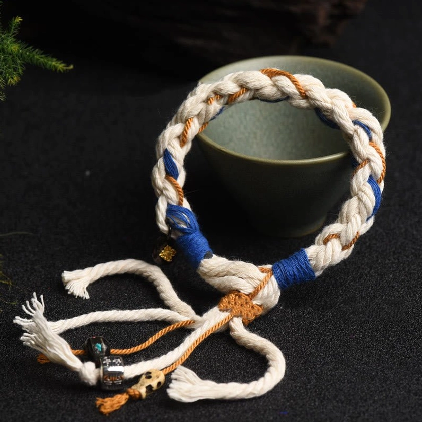 Close-up of handmade prosperity bracelet featuring white blue and tan woven design with hanging charms and frayed tassels near green ceramic bowl