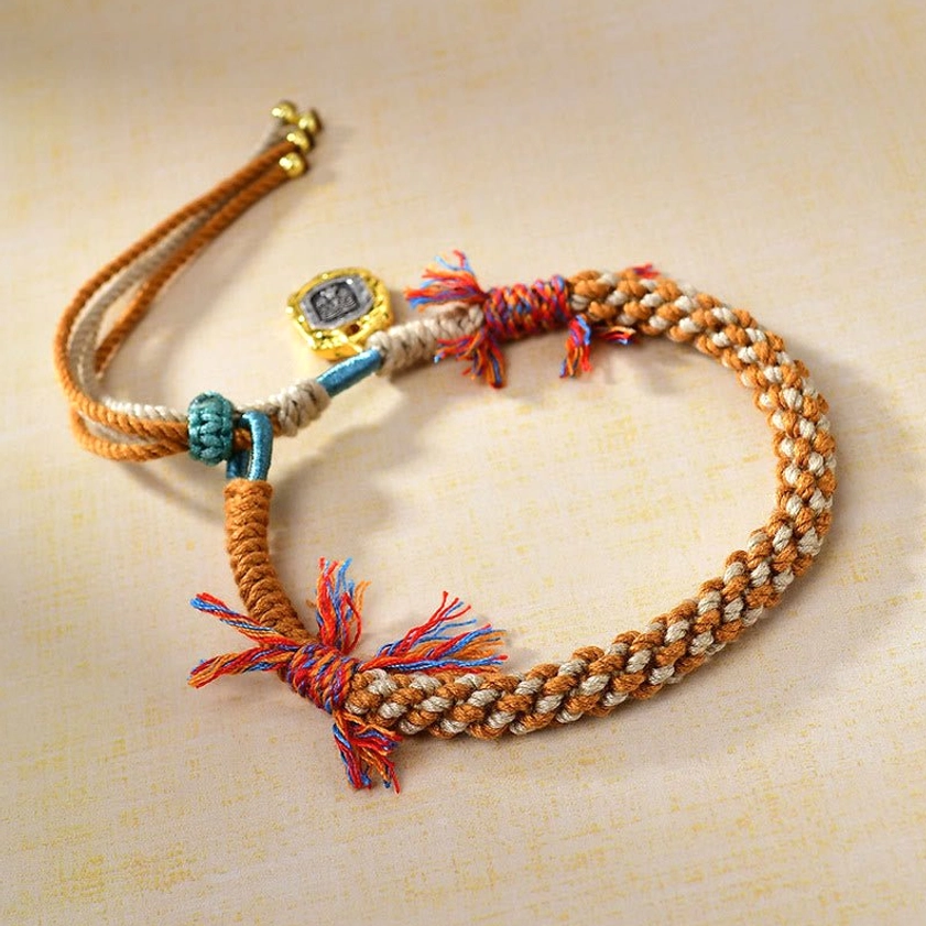 Top-down view of Buddhist wealth protection bracelet showcasing intricate braided multicolor rope pattern, dual-tone design, colorful tassels, and engraved square gold charm featuring Zakiram goddess symbol