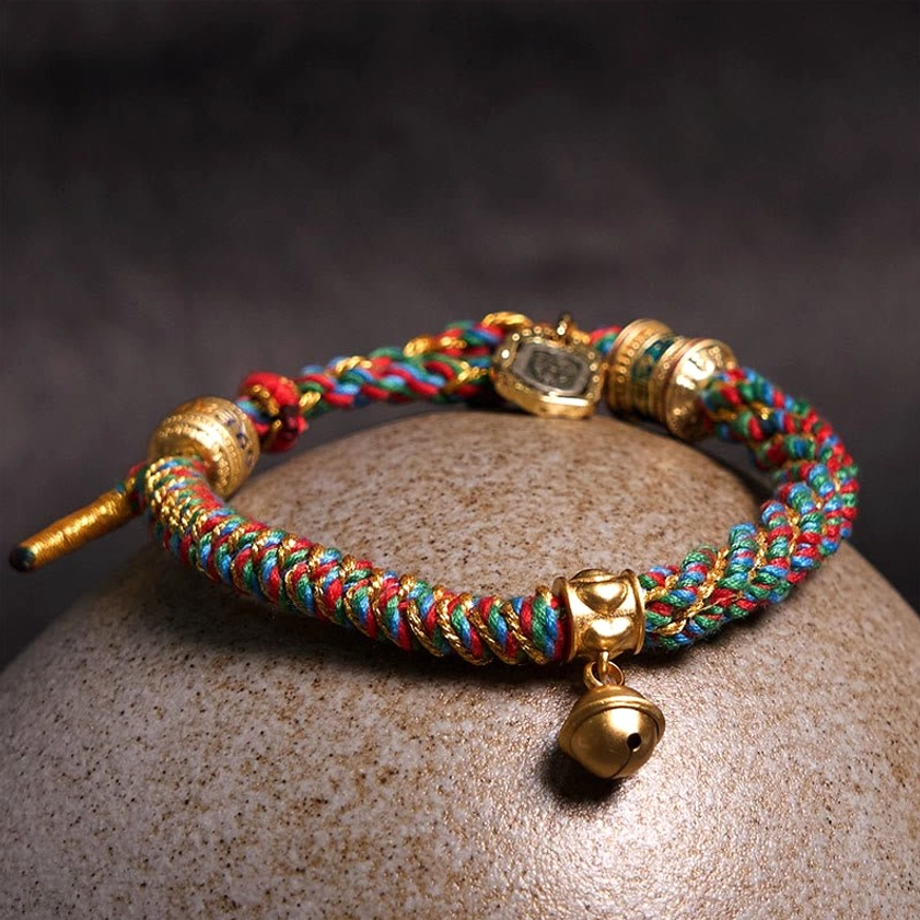 Top-down view of colorful woven cord Buddhist bracelet with gold hardware and hanging bell charm on stone surface