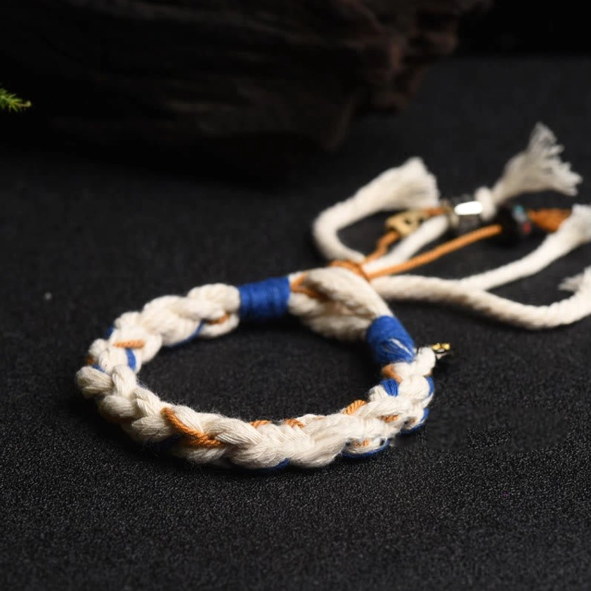 Tibetan style Zakiram bracelet on dark wooden textured board showing tri-color woven cord with blue wraps and frayed tassel details
