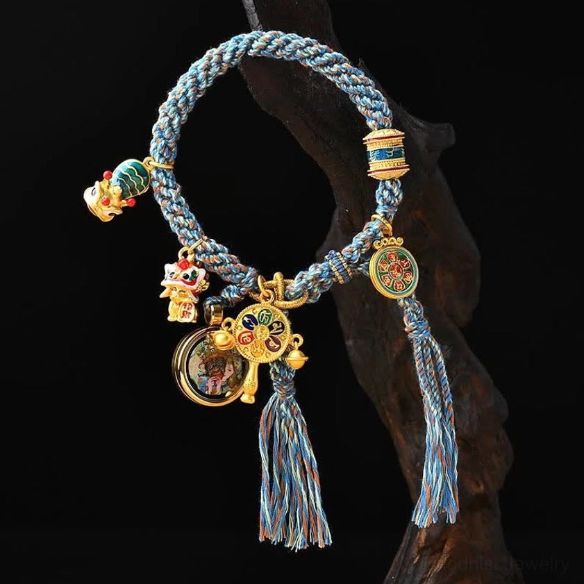 Zakiram Goddess of Wealth Buddhist blessing bracelet with blue braided string and gold metal charms hanging on dark wooden display