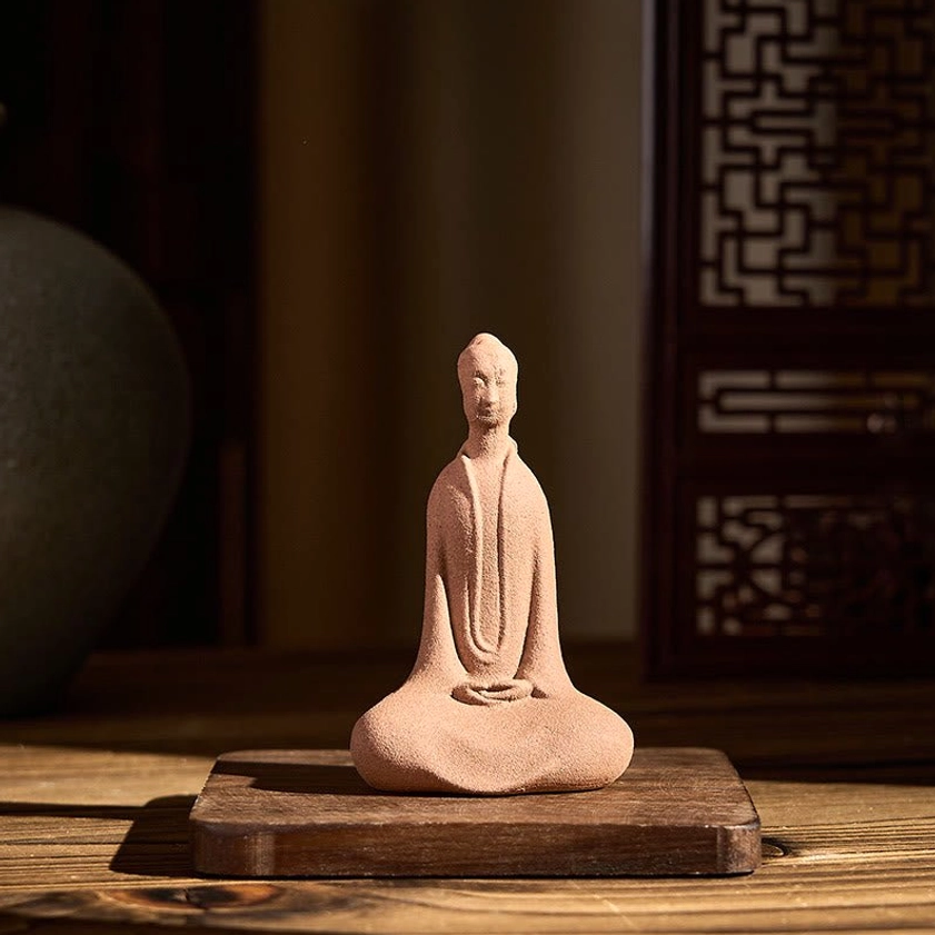 Buddha meditation statue with light earthy ceramic finish, cross-legged posture displayed on wooden base in traditional setting