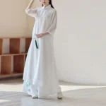 Zen meditation cheongsam dress with wide leg pants set, worn in three-quarter side view showing white sheer chiffon fabric and blue floral embroidery with green tassel pendant