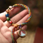 Chinese zodiac natal Buddha cinnabar blessing bracelet displayed on human palm, showing colorful braided silk rope with glazed beads and round metal Buddha pendant