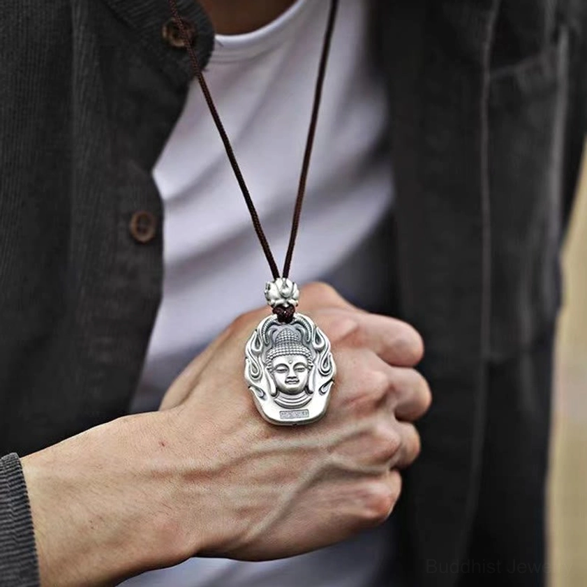 Buddhist pendant necklace worn on layered outfit, hand-wearing close-up showing dark brown cord, matte silver pendant against white shirt and dark coat